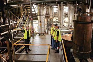 Four people wearing high visibility vests inside a warehouse with lots of pipes and a barrel.