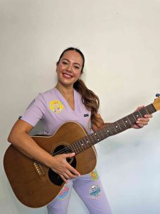 A woman wearing purple nursing scrubs holds a guitar
