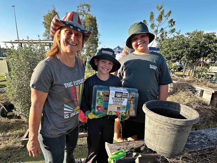 A woman and two boys wearing hats stand in front of a garden with pots and shovels
