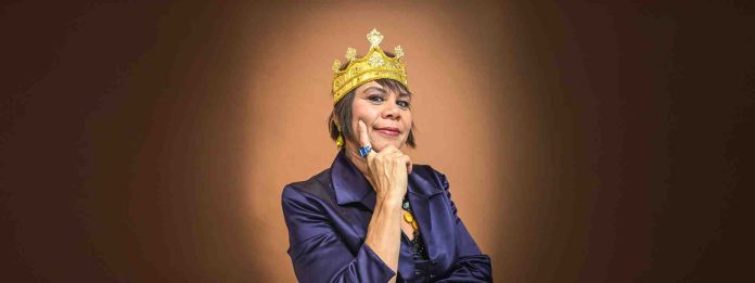 A woman wearing a purple jacket and a gold crown stands illuminated in front of a brown background.
